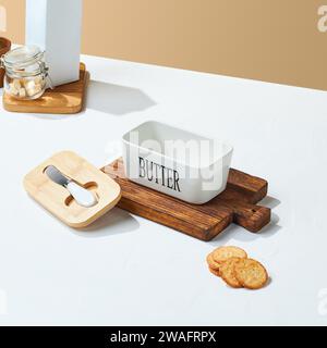 Weißer keramischer Butterbehälter mit Holzdeckel, auf einem Holztablett und Keks um ihn herum, auf einem weißen und cremefarbenen Hintergrund. Sauber Stockfoto