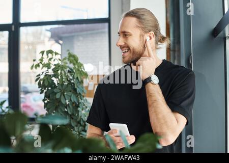 Fröhlicher Geschäftsmann in schwarzem T-Shirt, der Smartphone hält und Musik im Büro in Ohrhörern hört Stockfoto