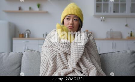 Gefrorene asiatische chinesische Frau in warmen Hut und Fäustlingen reiben gefrierende Hände sitzen in der kalten Küche Frosttemperatur Heizungsproblem in der Wohnung zu Hause Stockfoto