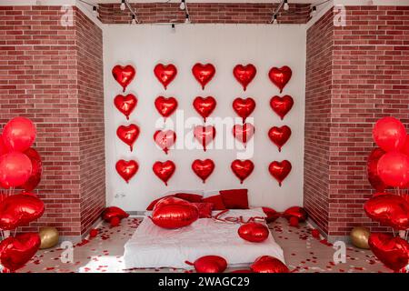 Weihnachtliches Loft-Interieur für Valentinstag. Ziegelwände weißes Bett auf dem Boden zwischen roten Herzballons mit der Inschrift Liebe, Kissen. Folie Stockfoto