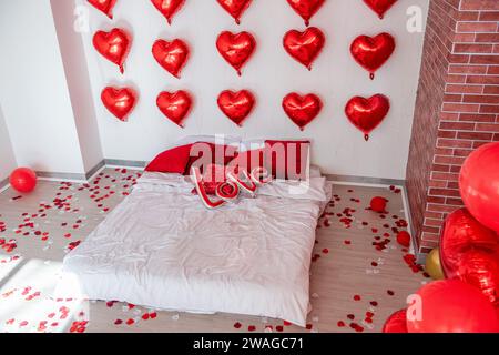 Weihnachtliches Loft-Interieur für Valentinstag. Ziegelwände weißes Bett auf dem Boden zwischen roten Herzballons mit der Inschrift Liebe, Kissen. Folie Stockfoto