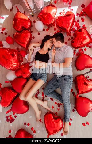 Blick von oben auf ein fröhliches junges Paar, das auf dem Boden in der Nähe eines roten weißen Ballons liegt, herzförmige Rosenblätter verstreut. Romantische Dekoration zum Valentinstag Stockfoto