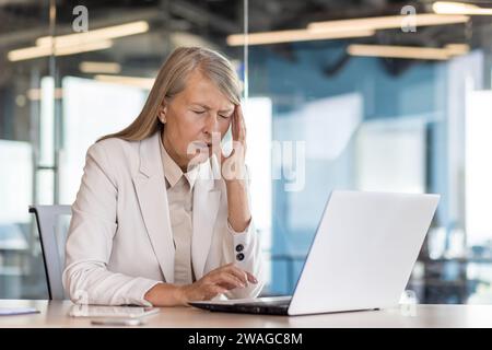 Ältere grauhaarige Geschäftsfrau, die im Büro arbeitet, am Schreibtisch sitzt, ihren Kopf mit der Hand hält und an Kopfschmerzen leidet. Stockfoto