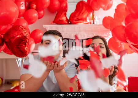 Nahaufnahme Porträt eines jungen Mannes und einer Frau, die nahe beieinander sitzen, vor einem weißen dekorativen Kamin, der mit roten Herzballons geschmückt ist. Paar Blo Stockfoto