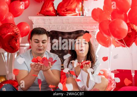 Nahaufnahme Porträt eines jungen Mannes und einer Frau, die nahe beieinander sitzen, vor einem weißen dekorativen Kamin, der mit roten Herzballons geschmückt ist. Paar Blo Stockfoto