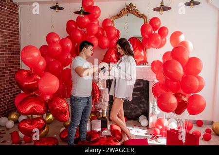 Fröhlicher Moment zwischen jungen Paaren, die mit Toast den Valentinstag in der Nähe von roten Ballons und weißem Kamin feiern. Frau lacht, hält Glas, kurz vor C Stockfoto