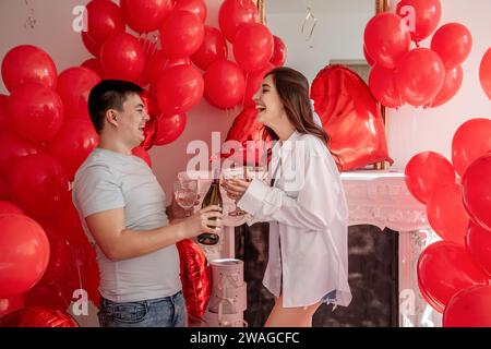 Fröhlicher Moment zwischen jungen Paaren, die mit Toast den Valentinstag in der Nähe von roten Ballons und weißem Kamin feiern. Frau lacht, hält Glas, kurz vor C Stockfoto