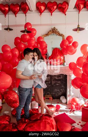 Fröhlicher Moment zwischen jungen Paaren, die mit Toast den Valentinstag in der Nähe von roten Ballons und weißem Kamin feiern. Frau lacht, hält Glas, kurz vor C Stockfoto