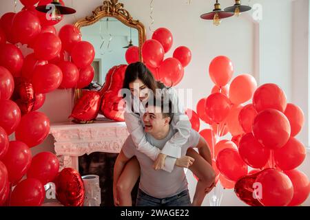 Verspielter, romantischer Moment zwischen verrücktem Paar in der Nähe von roten Ballons in einem hellen Raum mit weißer Inneneinrichtung. Frau sitzt auf dem Rücken des Mannes. Ein paar, das herumspielt Stockfoto