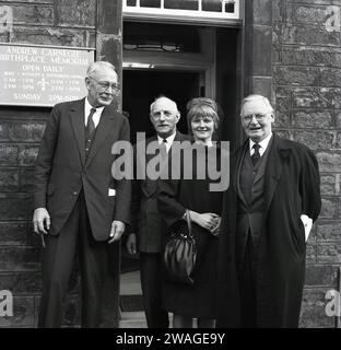 1960er Jahre, historisch, drei gut gekleidete ältere Männer und eine junge Dame stehen draußen am Eingang eines Steingebäudes mit einer Gedenktafel an der Wand, dem Andrew Carnegie Birthplace Memorial, täglich geöffnet. 1835 in einem Weberhaus in Moodie Street, Dunfermline, Fife, geboren, verließen Andrew Carnegie und seine Familie Schottland 1848, um ein neues Leben in Amerika zu führen. Das ursprüngliche Haus wurde 1908 für Besucher eröffnet, 1928 wurde der Museumsstandort um einen Gedenksaal erweitert. Stockfoto