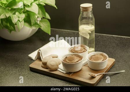 Schüsseln mit hausgemachtem Tiramisu-Dessert auf einem Holzbrett, Löffel mit Kakaopulver, Biskuitkuchen und einem Tuch Stockfoto