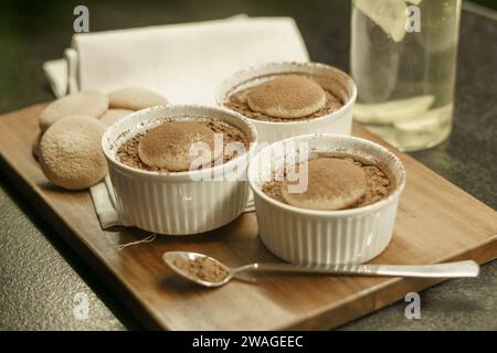 Schüsseln mit hausgemachtem Tiramisu-Dessert auf einem Holzbrett, Löffel mit Kakaopulver, Biskuitkuchen und einem Tuch Stockfoto