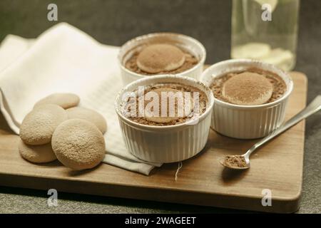 Schüsseln mit hausgemachtem Tiramisu-Dessert auf einem Holzbrett, Löffel mit Kakaopulver, Biskuitkuchen und einem Tuch Stockfoto