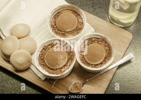 Schüsseln mit hausgemachtem Tiramisu-Dessert auf einem Holzbrett, Löffel mit Kakaopulver, Biskuitkuchen und einem Tuch Stockfoto