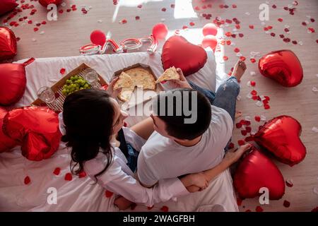 Blick von oben auf junge Paare, die sich gegenseitig füttern, Pizza aus der Take-away-Box genießen, mit Gläsern Champagner und grünen Trauben. Mann und Frau teilen sich festlich, Stockfoto