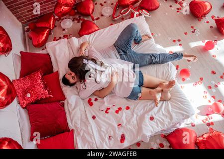 Verliebtes Paar umarmt sich eng auf weißem Bett zwischen roten Ballons, Kissen, Herzen, Rosenblüten. Mann und Frau sehen sich an, verbringen den Valentinstag zu Hause Stockfoto