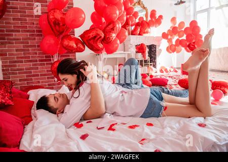 Verliebtes Paar umarmt sich eng auf weißem Bett zwischen roten Ballons, Kissen, Herzen, Rosenblüten. Mann und Frau sehen sich an, verbringen den Valentinstag zu Hause Stockfoto