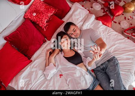 Blick von oben auf ein liebevolles Paar in legeren Kleidern auf weißem Bett mit roten Herzballons. Die Frau umarmt den Mann, sie sehen sich an, halten sich die Hände. Krieg Stockfoto