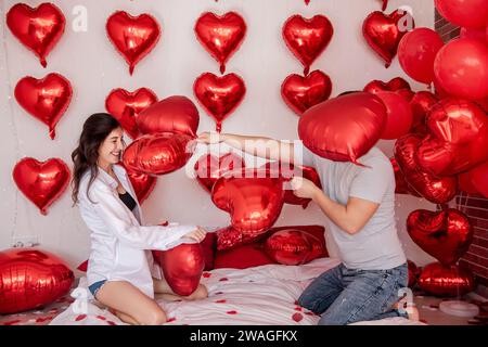 Verspielter Moment zwischen Paaren, die sich in Kissenballons auf dem Bett kämpfen, mit herzförmigen roten Ballons im Hintergrund. Die Frau macht fröhlich rumtäuscht Stockfoto