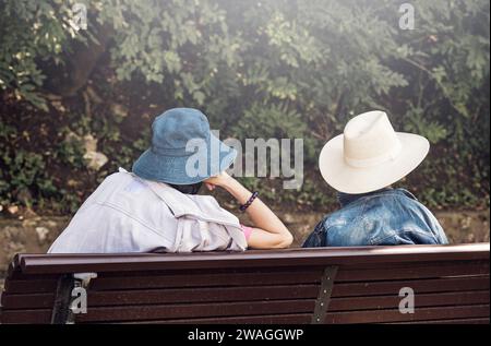Rückansicht mit zwei Frauen mit Sonnenhüten, die auf einer Bank im Park sitzen. Stockfoto