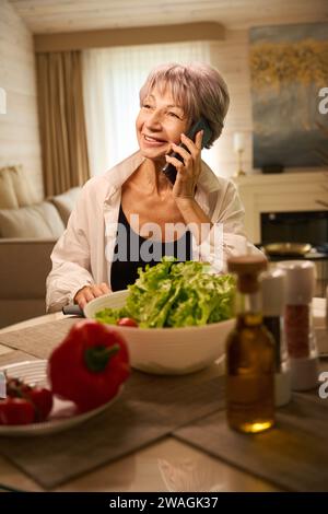 Nette ältere Dame kommuniziert über Handy in ihrer Küche Stockfoto
