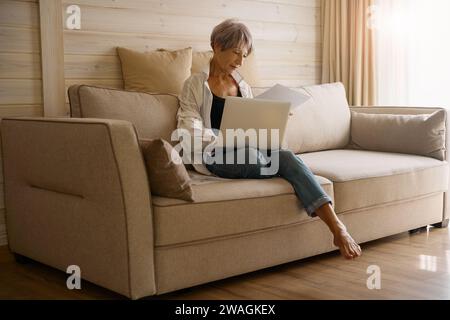 Ältere Frau mit kurzen Haarschnitten sitzt mit Laptop und Arbeitspapieren Stockfoto
