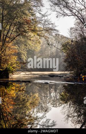 Eine ländliche Wasserlandschaft am Herbstmorgen mit Sonnenstrahlen im Hintergrund, die durch Bäume und buntes Laub leuchten Stockfoto