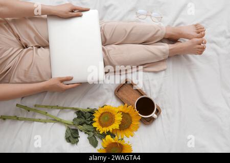 Frau mit modernem Laptop, einer Tasse Kaffee und wunderschönen Sonnenblumen im Bett Stockfoto