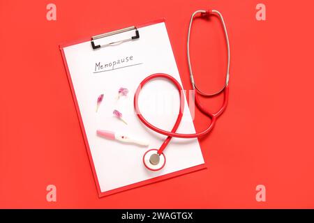 Clipboard mit Wort MENOPAUSE, Blumenblätter, Schwangerschaftstest und Stethoskop auf rotem Hintergrund Stockfoto