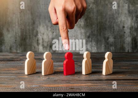 Eine menschliche Hand, die eine rote Holzfigur aus der Reihe der Holzfiguren auf einen Holztisch zeigt. Führungskonzept Stockfoto