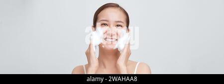 Glückliche Frau mit schäumendem Gesichtsreiniger waschendem Gesicht isoliert auf weißem Banner Hintergrund. Stockfoto