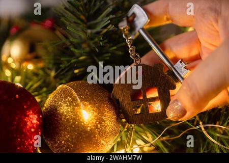 Geschenk Schlüsselhaus mit Schlüsselanhänger in der Hand Hintergrund des Weihnachtsbaums. Bauen, entwerfen, Projekt, Umzug in ein neues Haus, Hypothek, Miete und Kauf von Immobilien Stockfoto