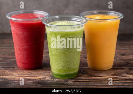 Drei verschiedene Frucht-Smoothies in Plastikgläsern zum Mitnehmen auf hölzernem Hintergrund Stockfoto