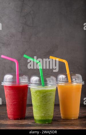 Drei verschiedene Frucht-Smoothies in Plastikgläsern zum Mitnehmen auf hölzernem Hintergrund Stockfoto