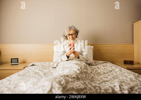 Eine ältere Frau sitzt morgens mit Pyjama auf dem Bett und liest ihre Nachrichten am Telefon. Stockfoto