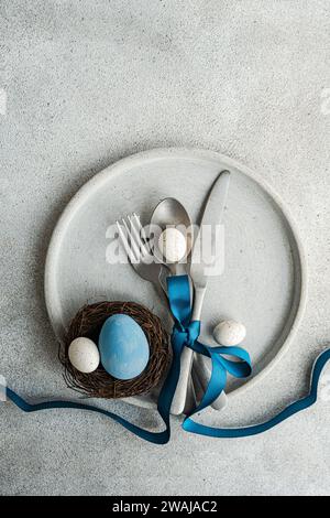 Blick von oben auf einen minimalistischen, aber stilvollen Ostertisch, mit Besteck, das mit blauem Band auf Betonplatte zusammengebunden ist, mit einem kleinen Nest mit Dekor Stockfoto
