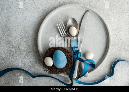 Blick von oben auf einen minimalistischen, aber stilvollen Ostertisch, mit Besteck, das mit blauem Band auf Betonplatte zusammengebunden ist, mit einem kleinen Nest mit Dekor Stockfoto