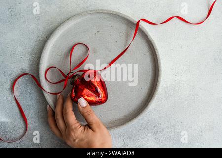 Von oben auf die Hand einer anonymen Frau, die ein rotes herzförmiges Ornament hält, gebunden mit einem lockigen roten Band, auf einer strukturierten Betonplatte auf grauem Sur platziert Stockfoto