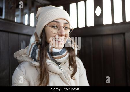 Porträt einer positiven jungen Frau in warmer Kleidung mit Halstuch und Beanie Hut lächelnd und in die Kamera schauend, während sie tagsüber in der Nähe eines Holzzauns stand Stockfoto