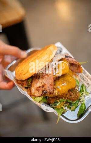 Von oben beschnittene nicht erkennbare Hand, die einen Gourmet-Beef-Burger mit Cheddar, knusprigem Speck, karamelisierten Zwiebeln und Gemüse auf einer Brioche hält Stockfoto