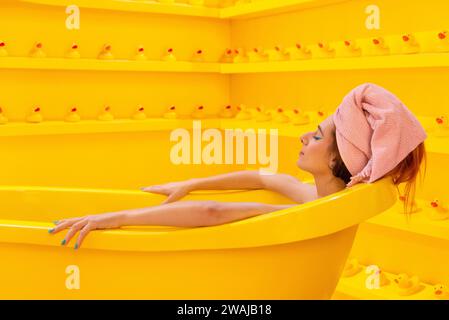 Frau, die sich in einer gelben Badewanne, umgeben von Gummienten, in einem monochromatischen gelben Zimmer entspannt Stockfoto