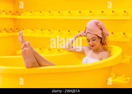 Zufriedene Frau mit einem Handtuch auf dem Kopf, das in einer leuchtend gelben Badewanne mit Gummienten im Hintergrund liegt Stockfoto