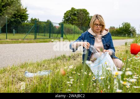 Eine konzentrierte blonde Frau in lässiger Kleidung mit einem weißen Müllsack, der auf dem Boden kniet und Plastikmüll aus dem Gras neben einem ländlichen Weg sammelt Stockfoto
