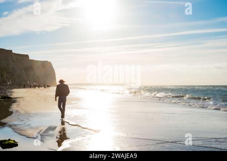 Dieses Bild fängt den ruhigen Moment einer einsamen Figur ein, die an einem sonnigen Strand entlang spaziert. Die Sonne hängt tief am Himmel und erzeugt ein warmes Leuchten und eine ruhige Atmosphäre. Die Figur ist mit dem hellen Licht versehen, das von den sanften Wellen reflektiert wird, und die Klippen im Hintergrund sorgen für ein Gefühl von Einsamkeit und Besinnung. Die Bildkomposition führt den Blick zum Horizont, wo das Meer in einem weichen Dunst auf den Himmel trifft und die Weite des Ozeans und die Einsamkeit des Individuums betont. Einsame Reflexion am Meer. Hochwertige Fotos Stockfoto