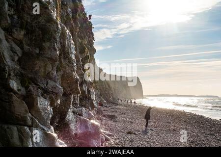 Ein beeindruckendes Bild, das eine Geschichte von Entdeckungen und Abenteuern entlang einer zerklüfteten Klippe erzählt. Der Fokus liegt hier auf der Figur, die parallel zur Felsenwand verläuft, die in Sonnenlicht getaucht ist und die komplizierten Details und Texturen der Felsen hervorhebt. Die Bewegung der Figur fügt dem Bild ein dynamisches Element hinzu, und die Anwesenheit anderer entfernter Individuen deutet auf einen beliebten und dennoch ruhigen Ort für Wanderungen am Meer hin. Der Kiesstrand unter den Füßen und das weitläufige Meer, das unter der hellen Sonne den Horizont trifft, runden diese Szene der natürlichen Majestät und menschlichen Neugier ab. Wanderlust An Den Klippen. Hohe Qualität p Stockfoto