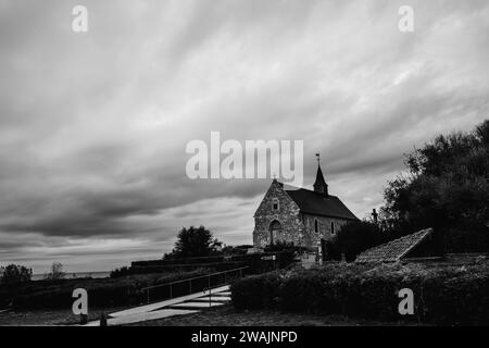 Dieses monochrome Bild zeigt eine Steinkirche in der Nähe der französischen Küste am Cap Blanc Nez. Die Kirche mit ihrer traditionellen Architektur steht vor einem dramatischen Himmel voller Textur und Bewegung, in Graustufen erfasst. Der Kontrast der dunklen Wolken und der hellen Steinfassade unterstreicht die dauerhafte Präsenz des Gebäudes. Das Bild erinnert an eine zeitlose Atmosphäre, die die historische Tiefe der Gegend und die Ruhe eines spirituellen Rückzugs am Meer widerspiegelt. Coastal Sanctuary: Steinkirche am Cap Blanc Nez. Hochwertige Fotos Stockfoto