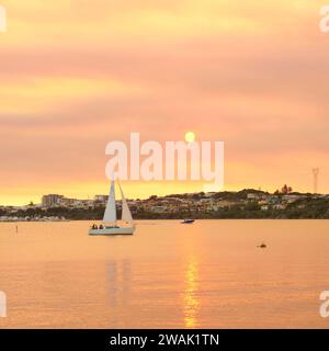 Quadratisches Bild einer Yacht und gelber und orangener Rauchnebel von einem vorgeschriebenen Brand, um Buschfeuer zu verhindern, Bicton, Swan River, Perth, Western Australia. Stockfoto