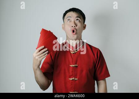 Schockiertes Gesicht eines asiatischen Mannes, der Cheongsam oder traditionelles chinesisches Tuch trägt, während er sich mit Angpao oder rotem Geldgeschenk auf weißem Hintergrund fächert Stockfoto