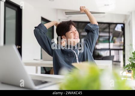 Junger asiatischer Mitarbeiter, der auf dem Stuhl sitzt und sich ausdehnt, um sich zu entspannen, während er hart im Büro arbeitet Stockfoto