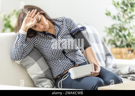 Eine junge Frau hat ein Gleichgewichtsproblem, sitzt auf dem Sofa und misst ihren Blutdruck mit einem digitalen Messgerät. Stockfoto